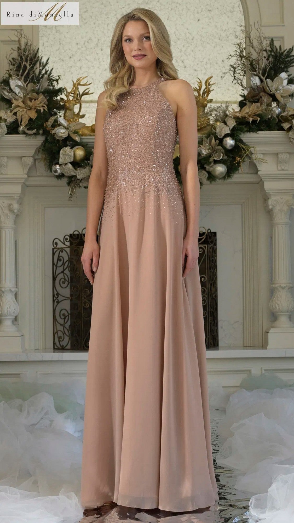 A woman stands indoors before a decorated mantelpiece wearing the Rina Di Montella RD3304, a rose gold sleeveless halter formal dress featuring a fitted beaded bodice and flowing chiffon A-line skirt.